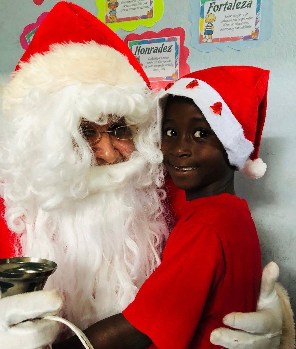 A boy in a Christmas hat hugs Santa Claus in the classroom
